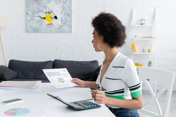 Mujer afroamericana mirando gráficos cerca de la calculadora y el teléfono inteligente en casa - foto de stock