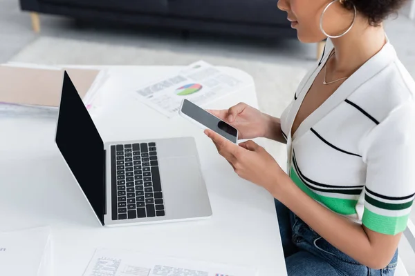 Vista cortada do freelancer afro-americano usando smartphone perto de laptop e papéis em casa — Fotografia de Stock