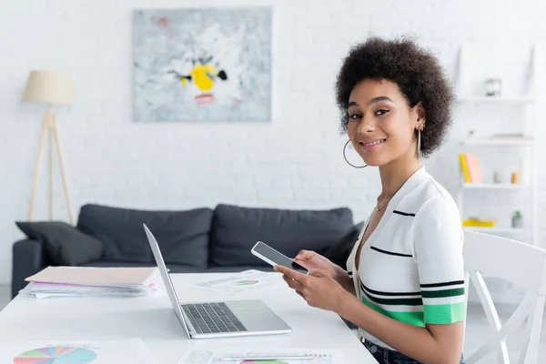 Alegre freelancer afroamericano sosteniendo teléfono inteligente cerca de la computadora portátil y gráficos en la mesa - foto de stock