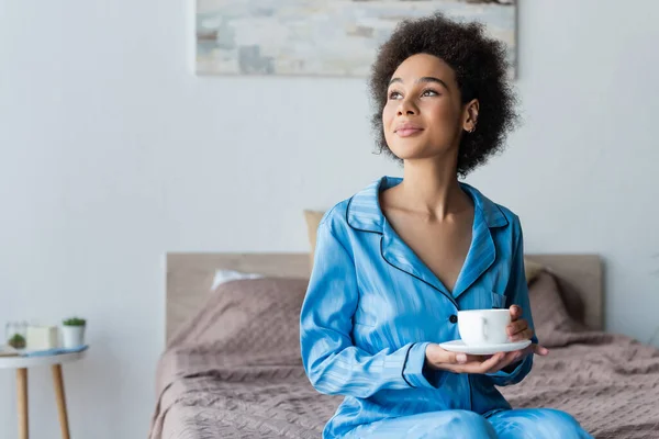 Joven mujer afroamericana en pijama sosteniendo taza blanca y platillo en el dormitorio - foto de stock