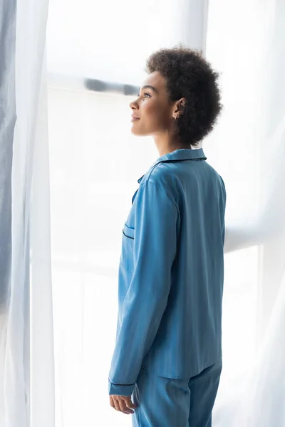 Vista lateral de mujer afroamericana en pijama azul de pie cerca de las cortinas - foto de stock