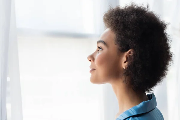 Vista laterale della donna afro-americana in pigiama in piedi vicino alla tenda a casa — Foto stock