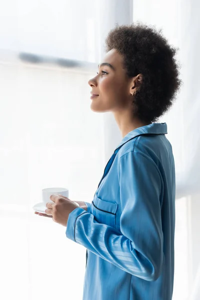 Vista lateral de mujer afroamericana en pijama sosteniendo taza cerca de cortinas en casa - foto de stock