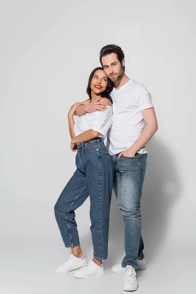 Ganzkörperansicht eines jungen Mannes, der eine brünette Frau umarmt, während er mit der Hand in einer Jeanstasche auf grau steht — Stockfoto