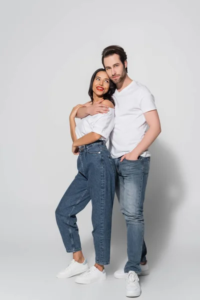 Vue pleine longueur du jeune homme embrassant femme brune tout en se tenant avec la main dans la poche de jeans sur gris — Photo de stock