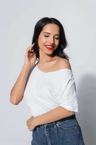 Sensual and smiling woman in white t-shirt with naked shoulder touching hair while posing on grey — Stock Photo