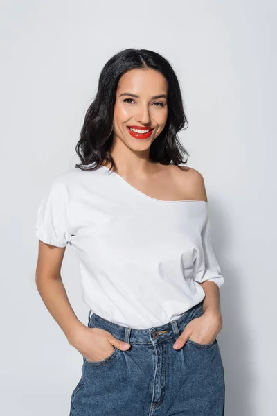 Mujer feliz con labios rojos y hombro desnudo posando con las manos en bolsillos de jeans en gris - foto de stock