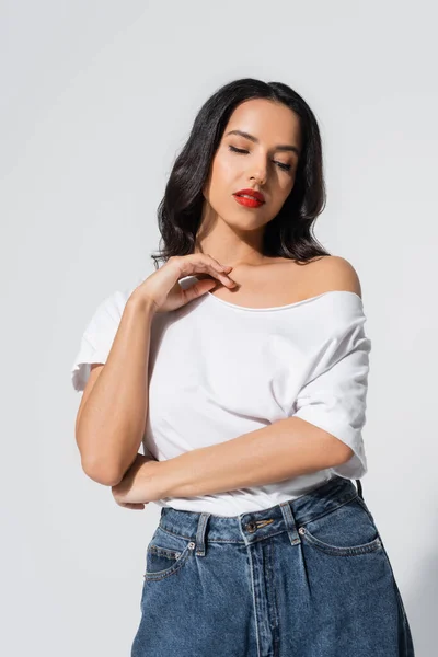 Joven morena en camiseta blanca con hombro desnudo posando sobre gris - foto de stock