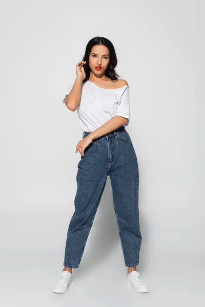 Vista completa de mujer morena en jeans y camiseta blanca posando sobre gris - foto de stock