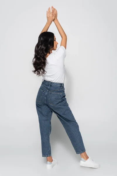 Full length view of woman in white t-shirt and jeans standing with raised hands on grey — Stock Photo