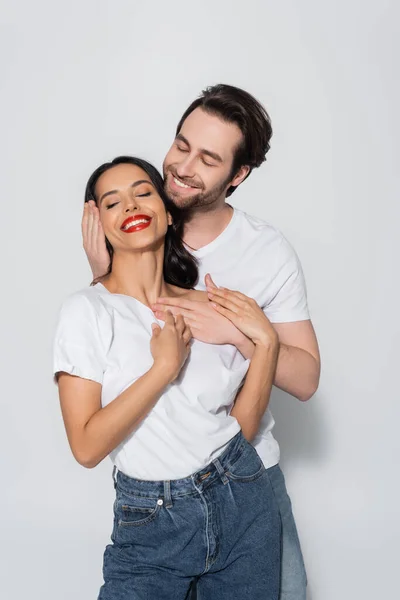 Sonriente hombre abrazando feliz morena mujer de pie con los ojos cerrados en gris - foto de stock