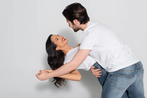 Jovem homem de t-shirt branca dançando com mulher morena sorridente em cinza — Fotografia de Stock