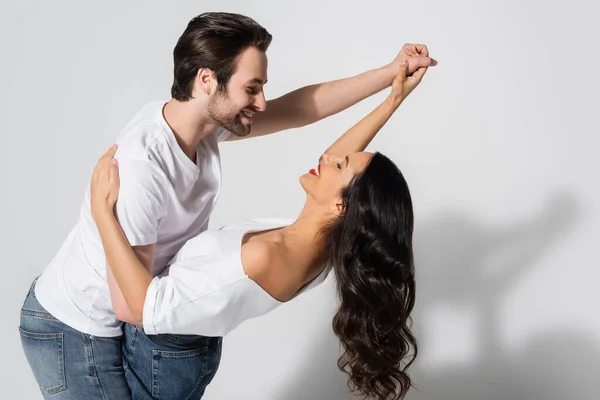 Smiling couple in jeans and white t-shirts smiling at each other while dancing on grey — Stock Photo