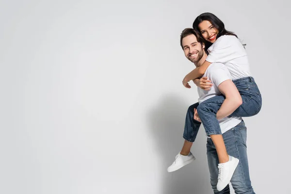 Alegre joven pareja en blanco camisetas y jeans mirando a la cámara mientras se divierten en gris - foto de stock