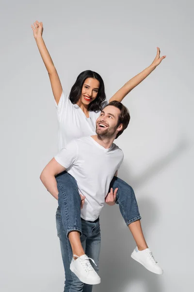 Joyeux homme piggyback excité brunette femme en t-shirt blanc et jeans sur gris — Photo de stock