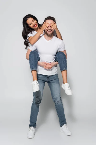 Joven hombre piggybacking alegre morena mujer cubriendo sus ojos en gris - foto de stock