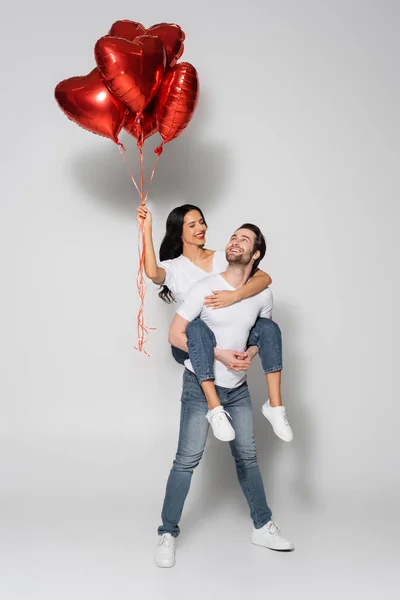 Piena lunghezza vista di felice uomo piggybacking fidanzata con palloncini a forma di cuore su grigio — Foto stock