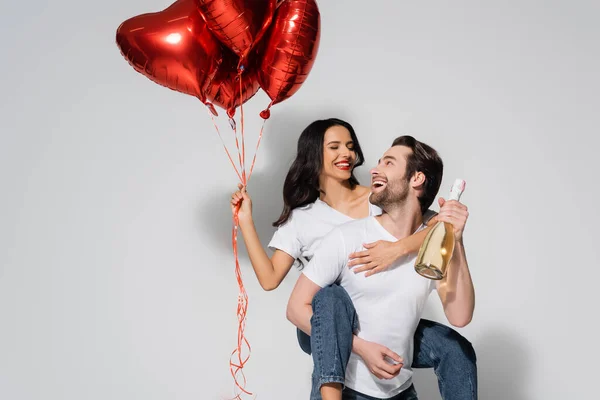 Homem segurando garrafa de champanhe enquanto piggybacking mulher alegre com balões em forma de coração no cinza — Fotografia de Stock