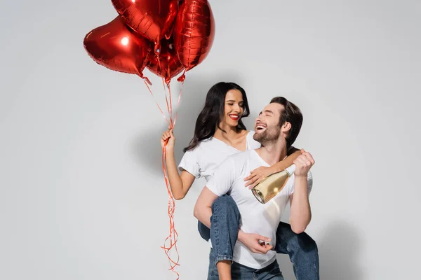 Alegre hombre con champán botella piggybacking mujer con rojo en forma de corazón globos en gris - foto de stock
