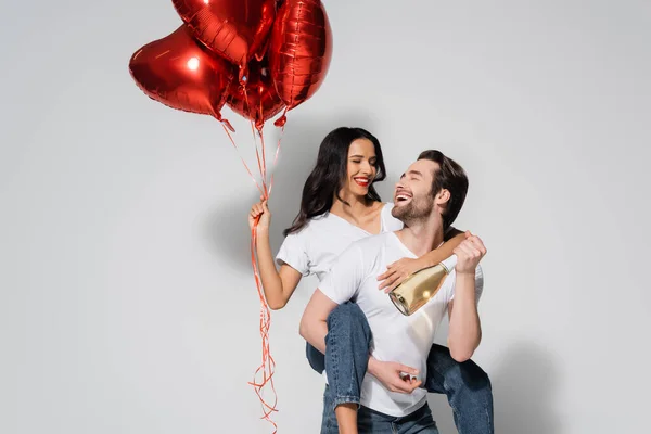 Allegro uomo con bottiglia di champagne a cavalletto donna con palloncini rossi a forma di cuore su grigio — Foto stock