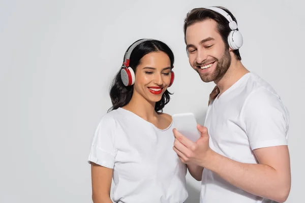 Joyful couple in white t-shirts and headphones looking at mobile phone on grey — Stock Photo