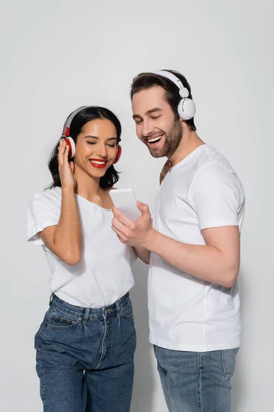 Homme souriant montrant smartphone à petite amie réglage casque sur gris — Photo de stock