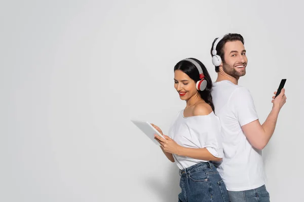 Feliz jovem casal em fones de ouvido usando dispositivos enquanto em pé de volta para trás no cinza — Fotografia de Stock