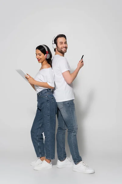 Visão comprimento total do casal alegre com fones de ouvido e gadgets de pé de volta para trás no cinza — Fotografia de Stock