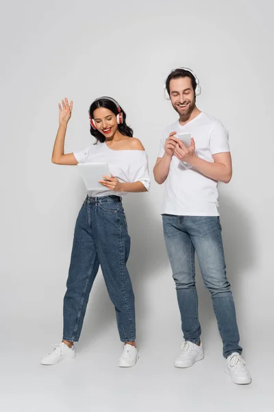 Mujer feliz en los auriculares saludando la mano durante la videollamada cerca de novio en gris - foto de stock