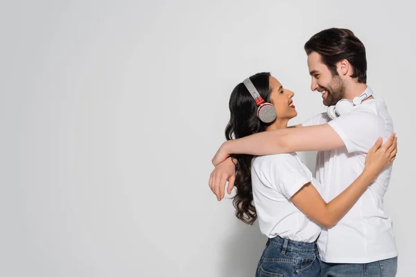 Vista lateral da mulher alegre em fones de ouvido perto namorado abraçando-a em cinza — Fotografia de Stock