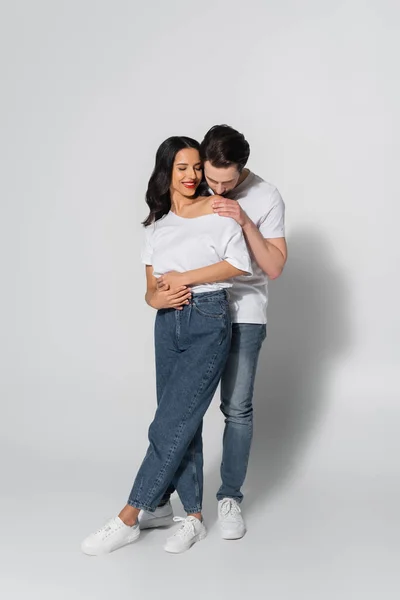 Full length view of young man kissing naked shoulder of woman wearing white t-shirt and jeans on grey — Stock Photo