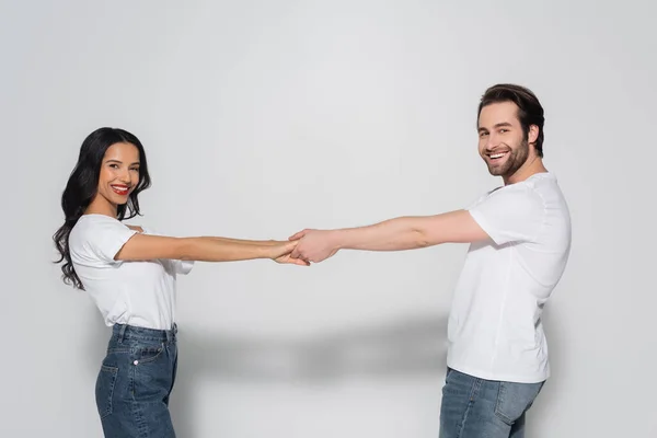 Casal alegre em camisetas brancas olhando para a câmera enquanto segurando as mãos no cinza — Stock Photo