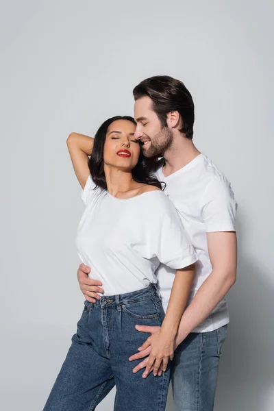 Улыбающийся мужчина обнимает соблазнительную женщину, стоящую с рукой за головой и закрытыми глазами на сером — стоковое фото
