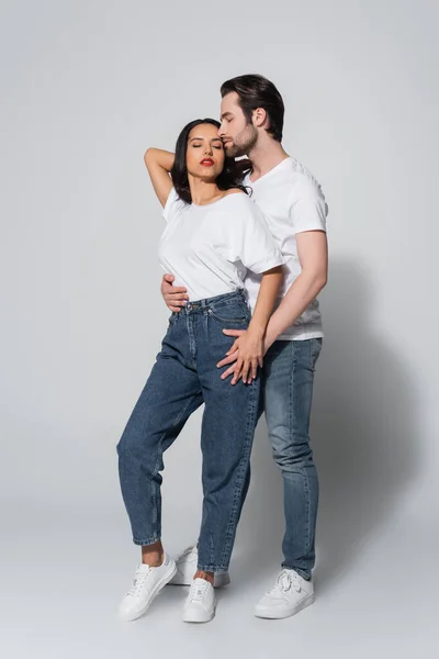 Vista a figura intera dell'uomo ad occhi chiusi che abbraccia seducente donna in t-shirt bianca e jeans su grigio — Foto stock