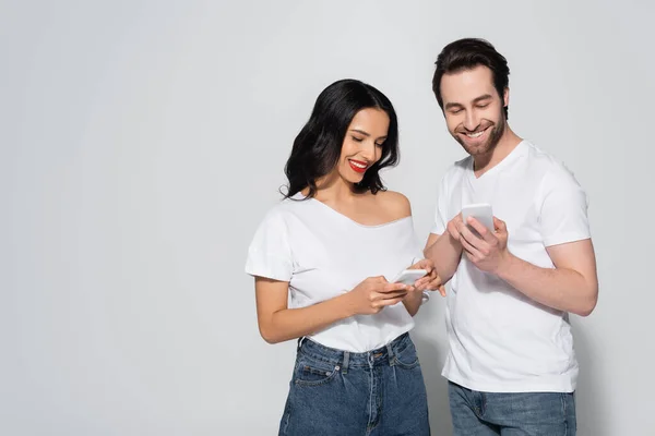 Mujer morena sonriente y hombre barbudo en camisetas blancas usando teléfonos inteligentes en gris - foto de stock