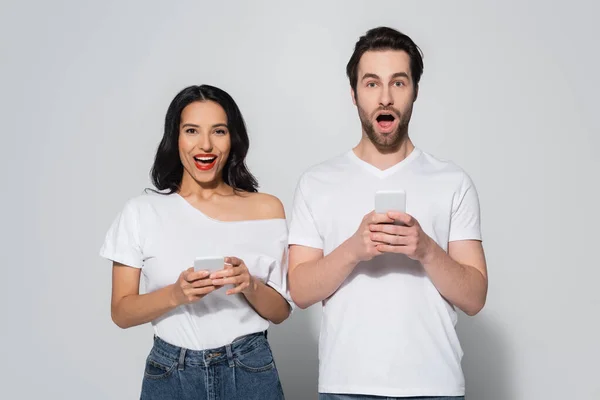 Здивована пара в білих футболках дивиться на камеру, використовуючи смартфони на сірому — стокове фото