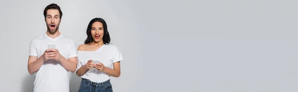 Astonished couple in casual clothes looking at camera while using smartphones on grey, banner — Stock Photo