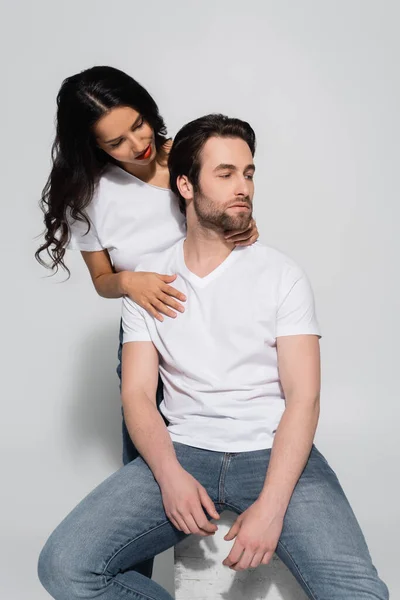 Sensual morena mujer abrazando al hombre en camiseta blanca y jeans sentados en gris - foto de stock