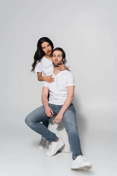 Hombre en jeans y camiseta blanca mirando a la cámara mientras está sentado cerca de morena seductora mujer en gris - foto de stock