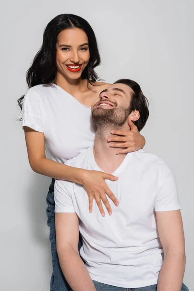 Femme séduisante regardant la caméra tout en embrassant jeune homme en t-shirt blanc sur gris — Photo de stock