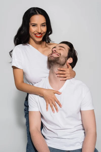 Mujer seductora mirando a la cámara mientras abraza joven en camiseta blanca en gris - foto de stock