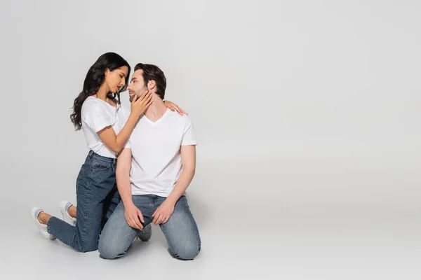 Vue pleine longueur de la femme passionnée touchant le visage du jeune homme tout en se tenant sur les genoux sur fond gris — Photo de stock