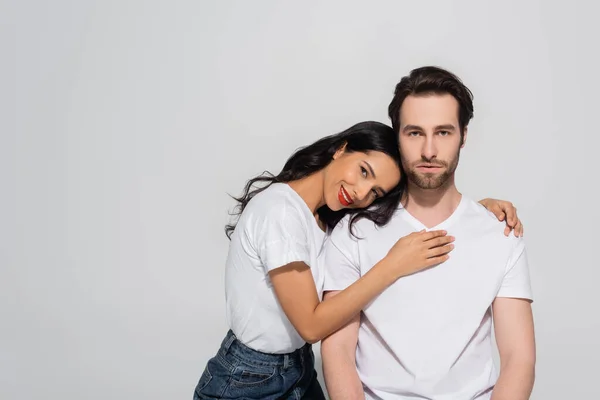 Mujer morena feliz en camiseta blanca apoyada en el novio mientras mira la cámara aislada en gris - foto de stock