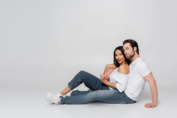 Vue pleine longueur du jeune couple en t-shirts et jeans blancs regardant la caméra assis sur le gris — Photo de stock