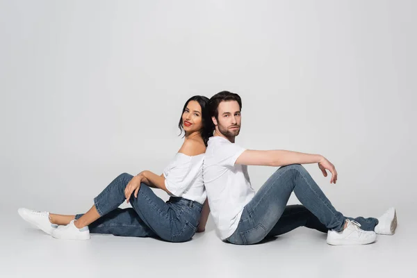 Vista de comprimento total do jovem casal em camisetas brancas e jeans sentado de volta para trás no cinza — Fotografia de Stock