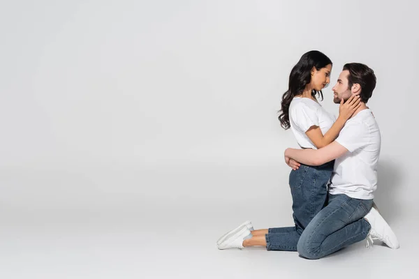 Vista lateral do casal em camisetas brancas e jeans de pé sobre os joelhos e abraçando em cinza — Fotografia de Stock