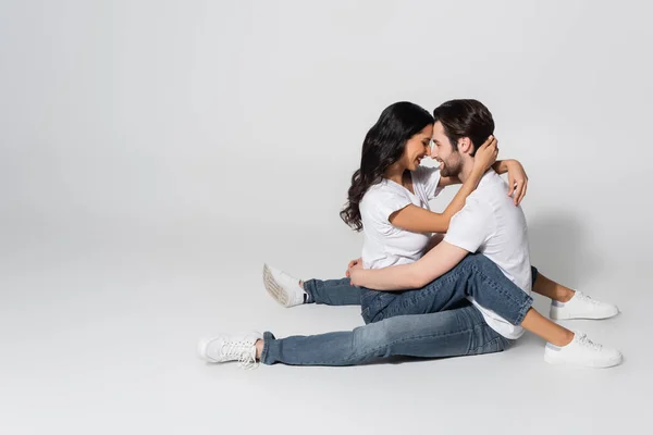 Young couple in white t-shirts and jeans embracing while sitting face to face on grey — Stock Photo