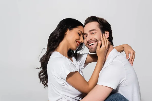 Cheerful man looking at camera near sexy brunette woman hugging his face isolated on grey — Stock Photo