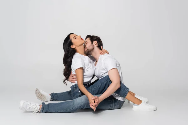 Homem de t-shirt branca e jeans de mãos dadas com mulher sedutora e beijá-la enquanto sentado em cinza — Fotografia de Stock