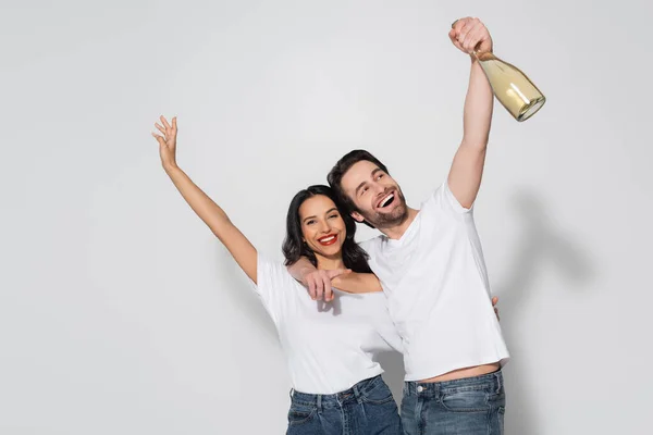Uomo allegro che tiene bottiglia di champagne e punta con il dito mentre abbraccia la ragazza sul grigio — Foto stock